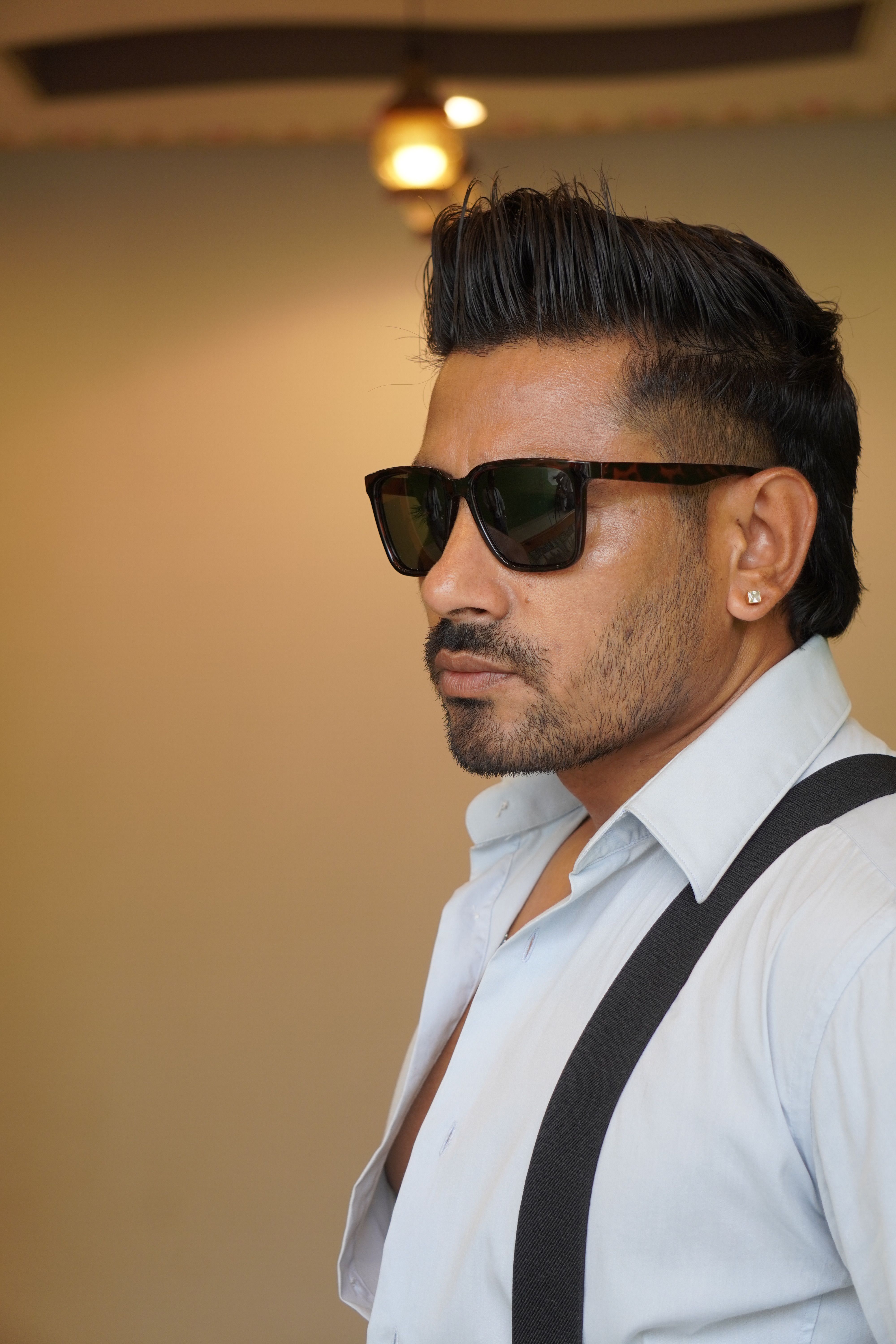Portrait of a man wearing sunglasses, shirt, and suspenders indoors.