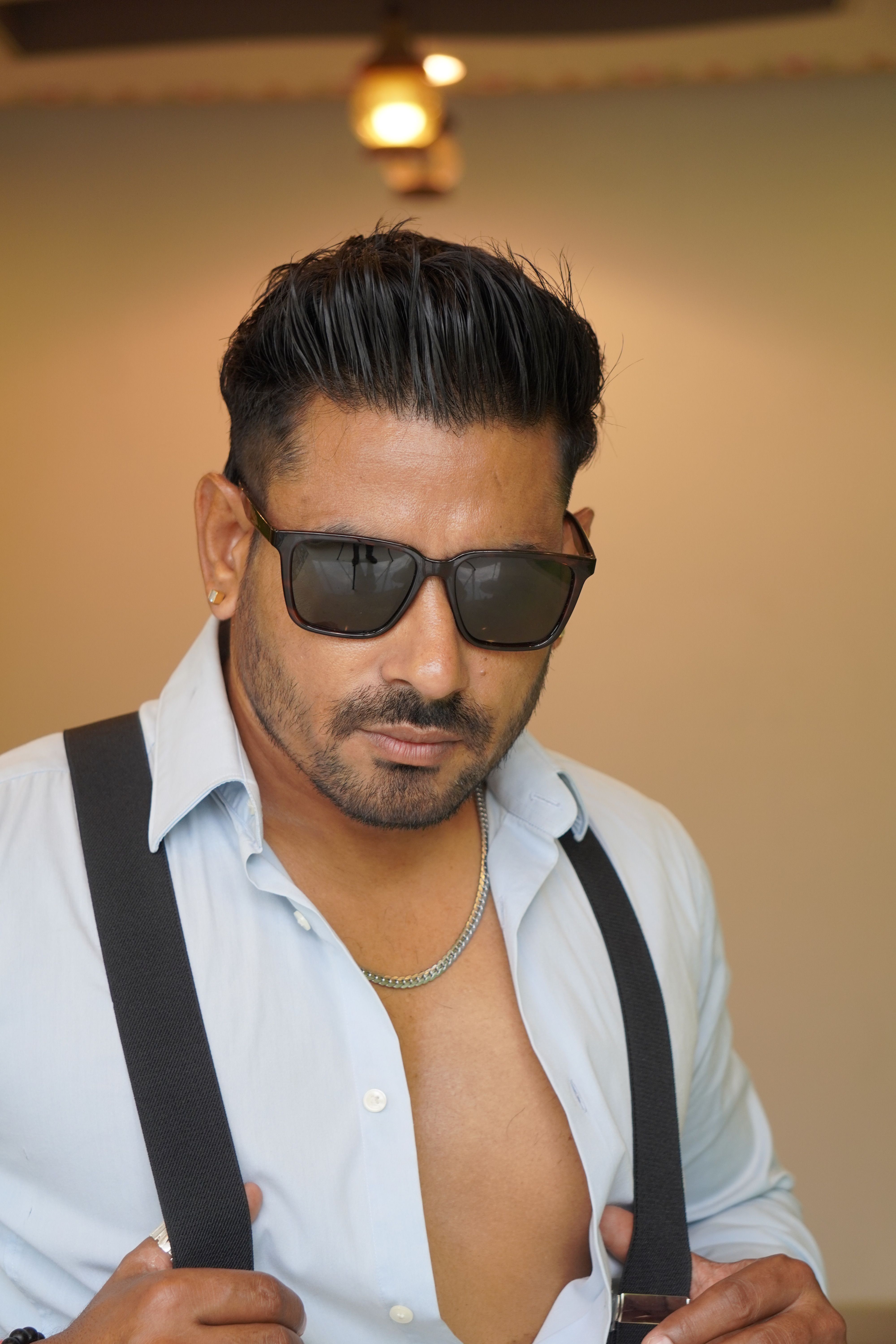 Stylish man wearing sunglasses, open shirt, and suspenders showing confidence indoors.