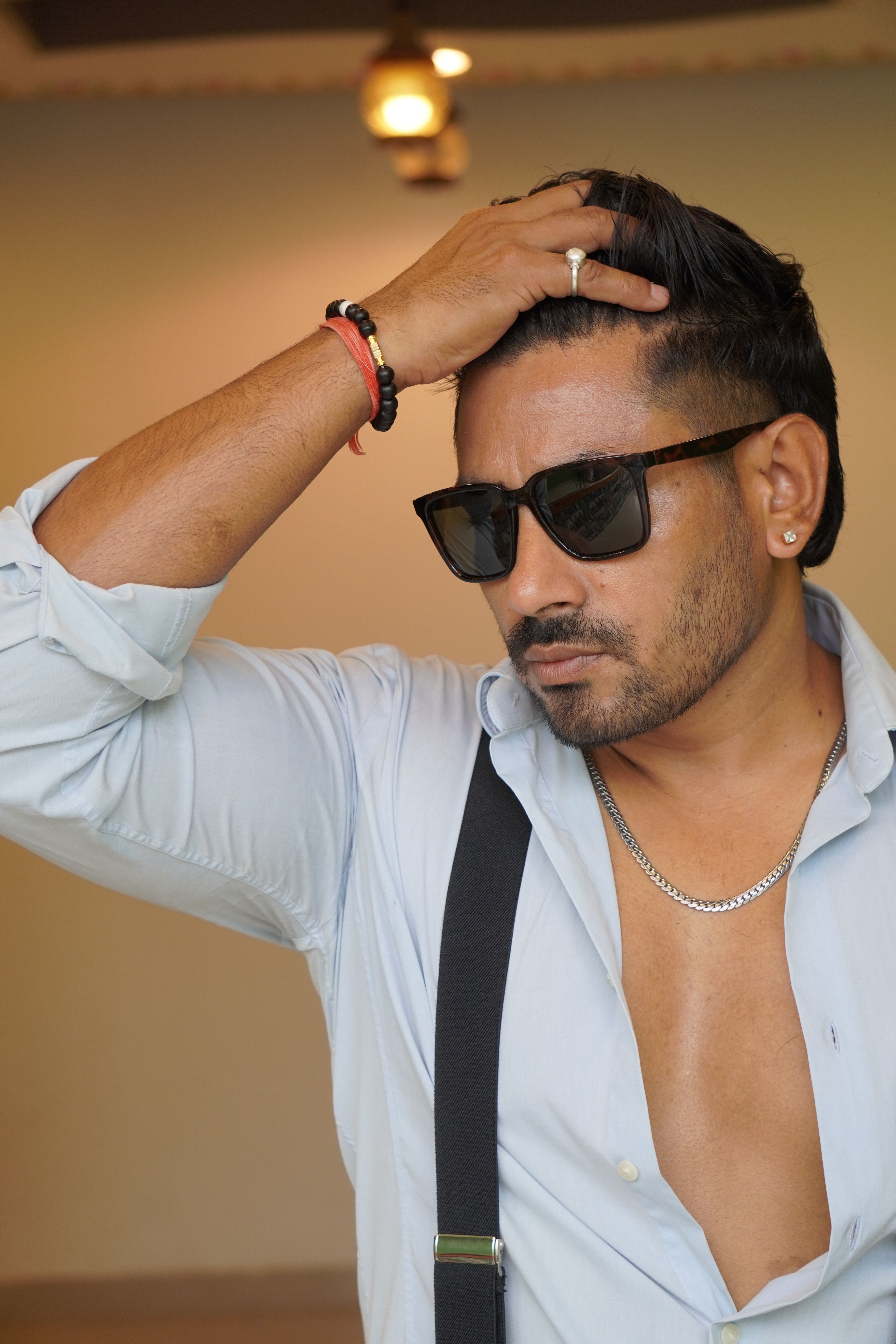 Portrait of a confident man wearing sunglasses, suspenders, and an open shirt indoors.
