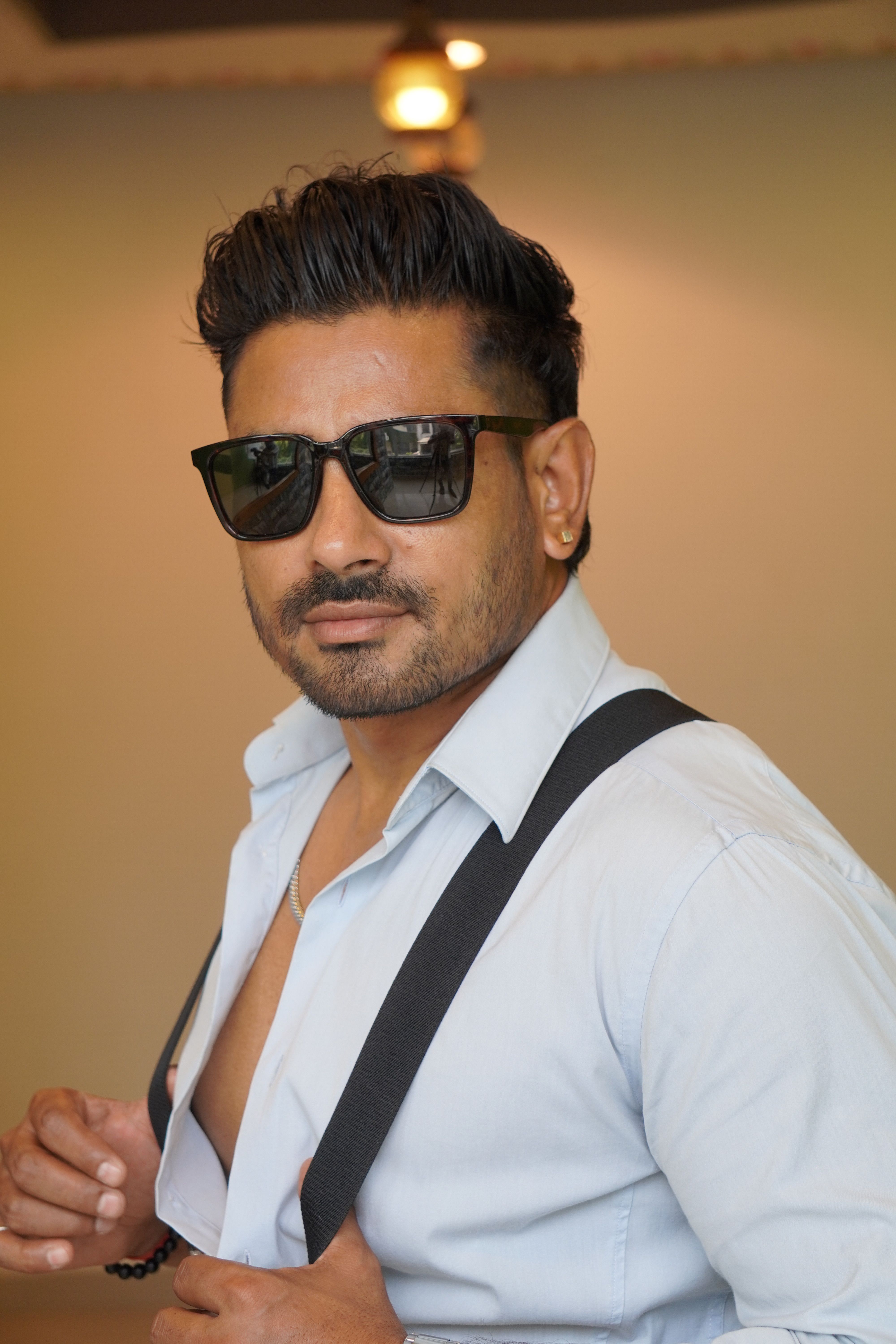 Portrait of a stylish young man with sunglasses and suspenders indoors, exuding confidence.