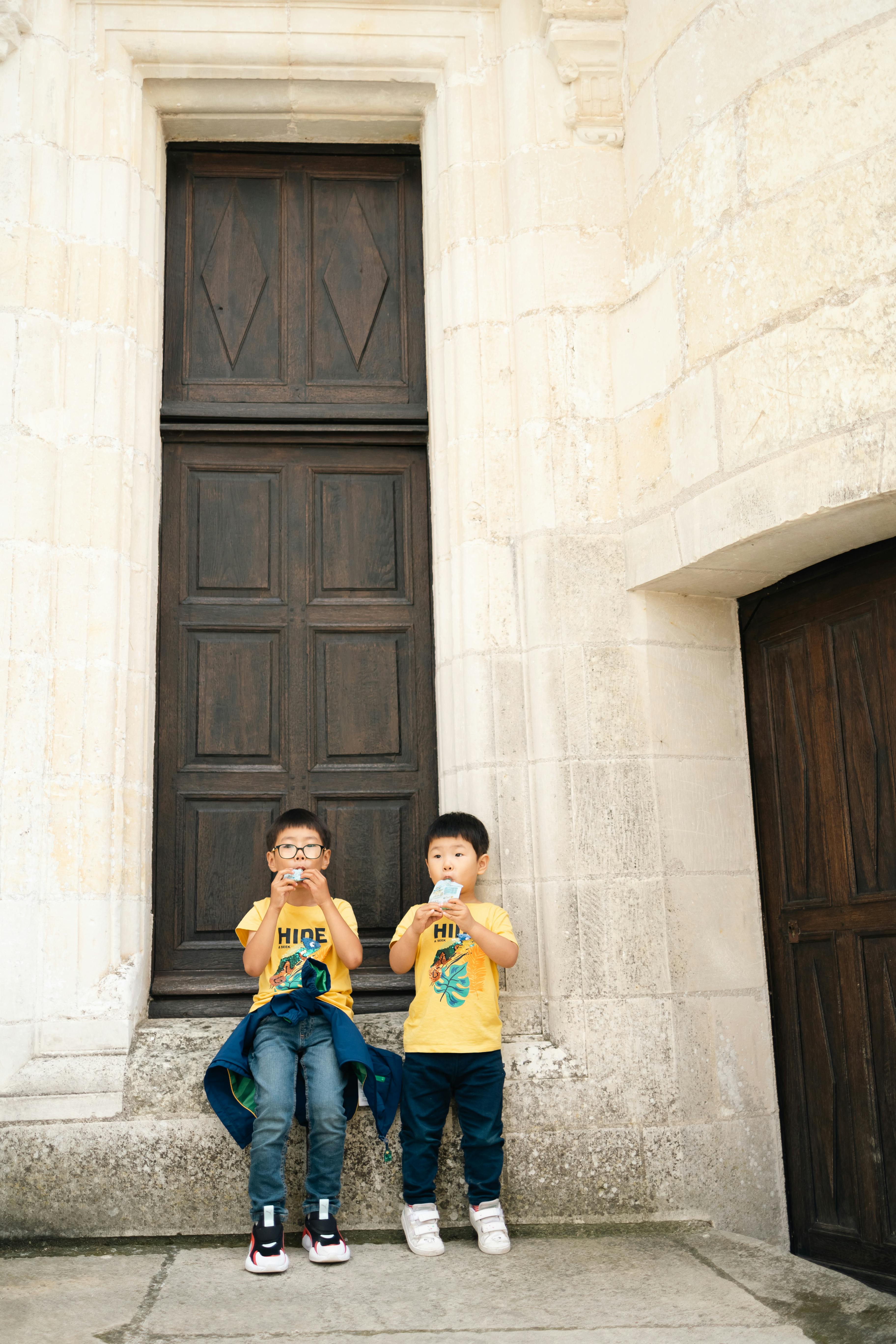 Two young boys in yellow shirts drinking juice in front of a large wooden door.