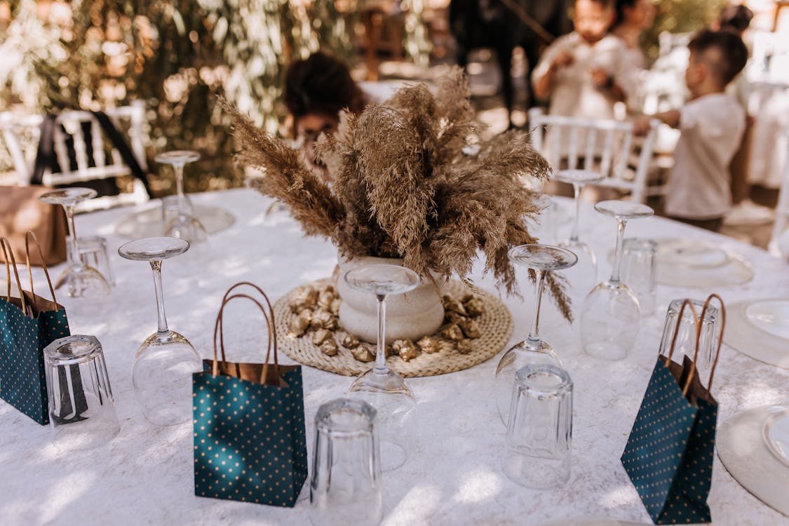 Outdoor wedding table setup featuring a rustic centerpiece with dried plants and gifts in teal dotted bags.