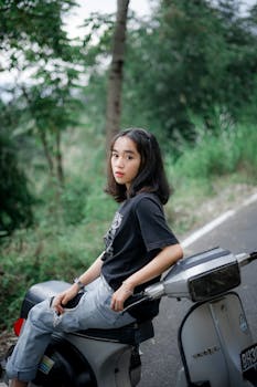 Asian woman sitting on scooter by the roadside in a lush, green landscape.