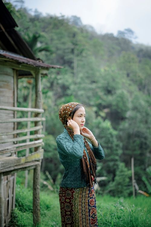 Free Asian woman wearing shawl stands by a rustic house in lush greenery. Stock Photo