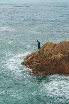 A lone fisherman stands on a rocky cliff with fishing rod, overlooking the vast ocean.