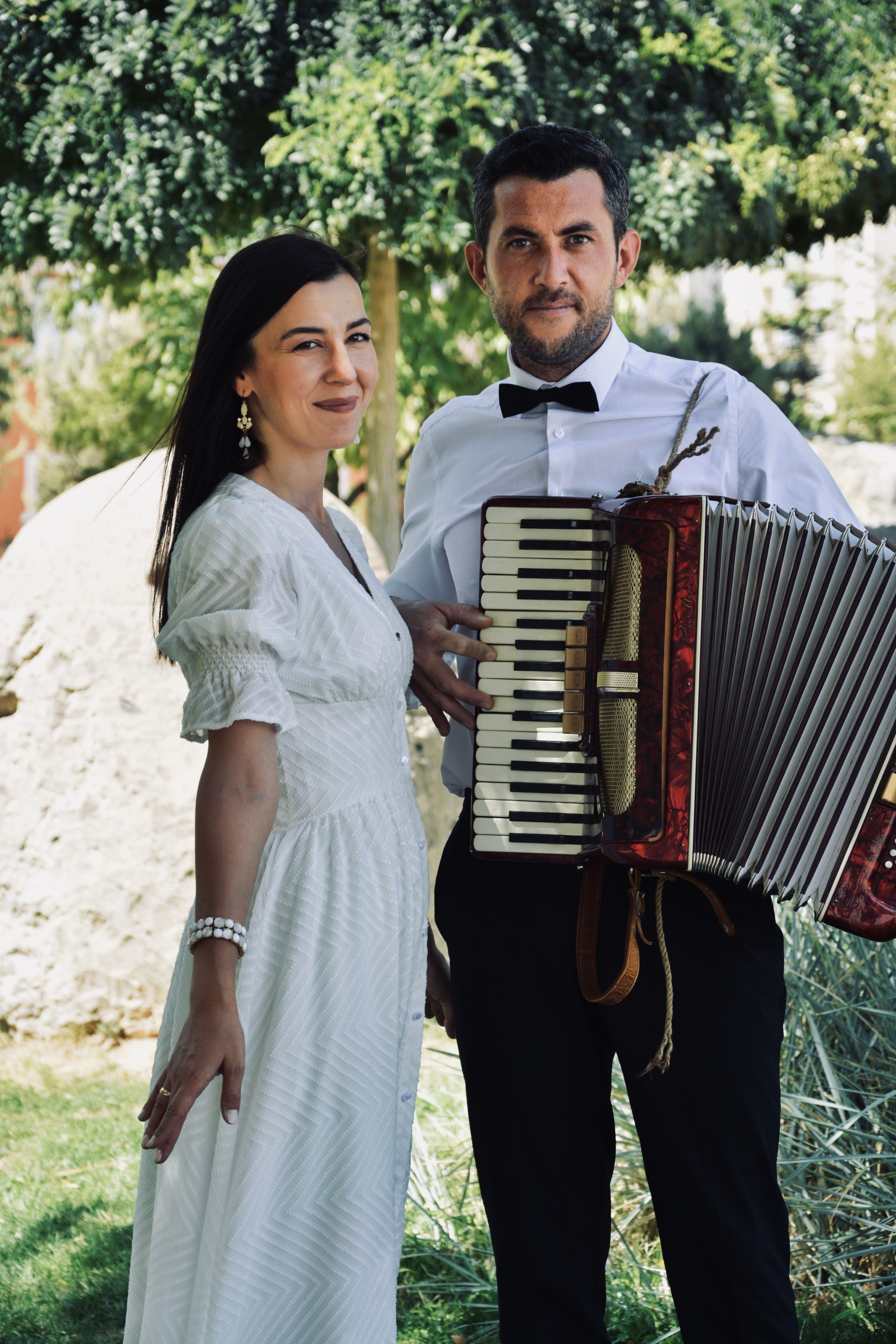 A man playing an accordion with a woman beside him in a sunny outdoor setting.