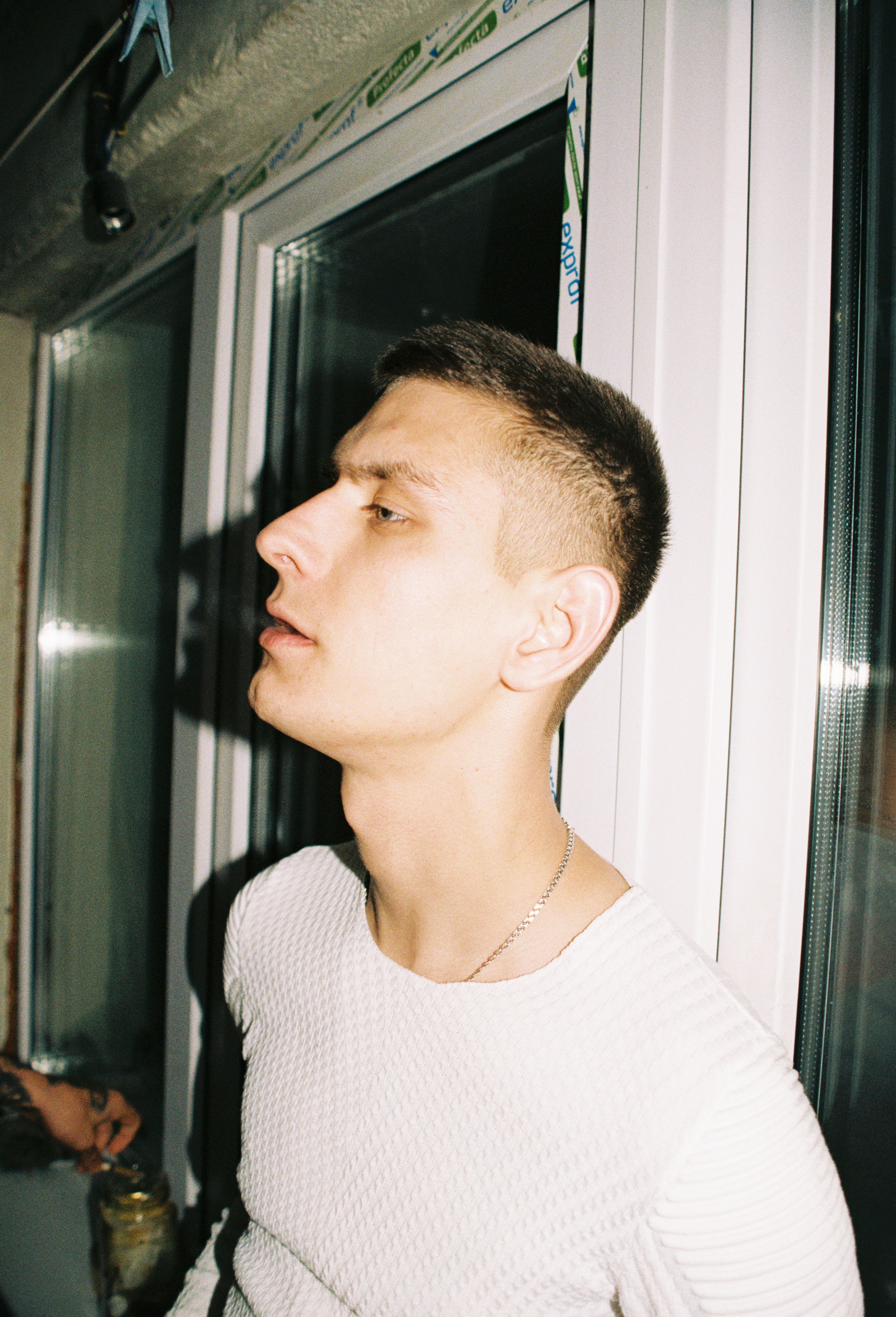 Profile portrait of young man in white sweater standing by a window indoors.