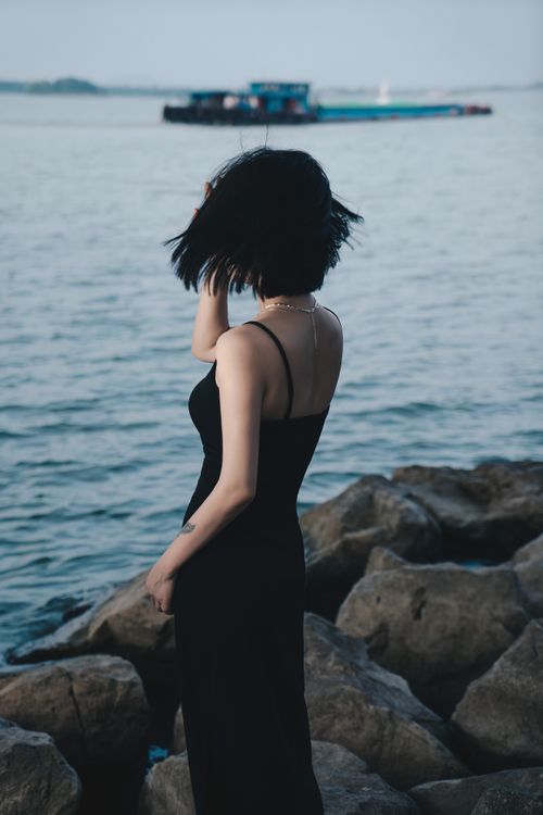 Free A woman in a black dress stands on rocky shoreline overlooking the sea. Stock Photo