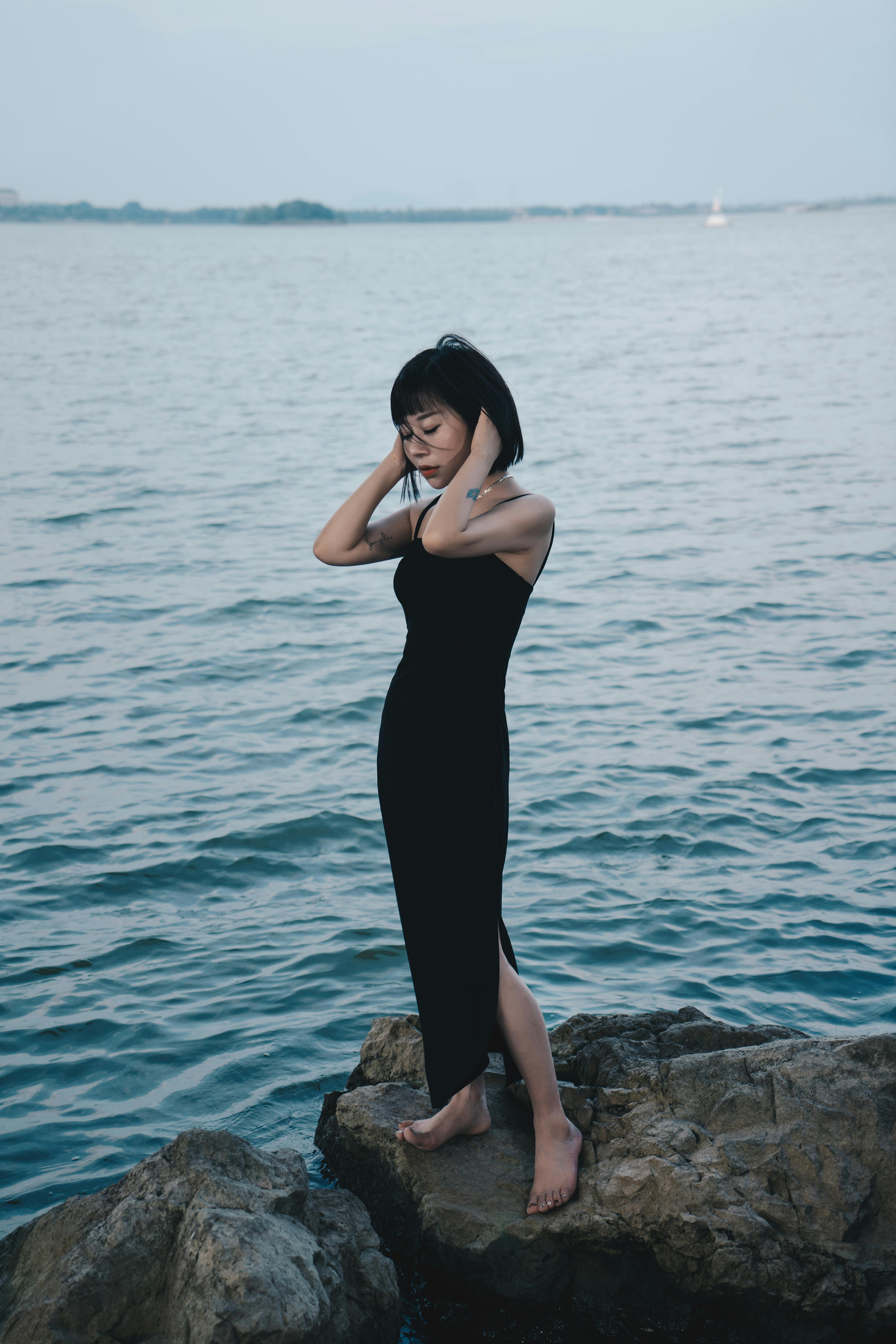 A woman in a black dress poses on rocky seaside, evoking calm and elegance.