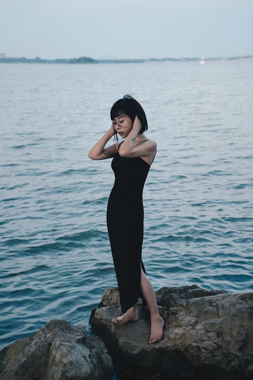 Free Young Woman in a Long Sleeveless Pencil Dress Standing Barefoot on Wet Rocks at the Sea Shore Stock Photo
