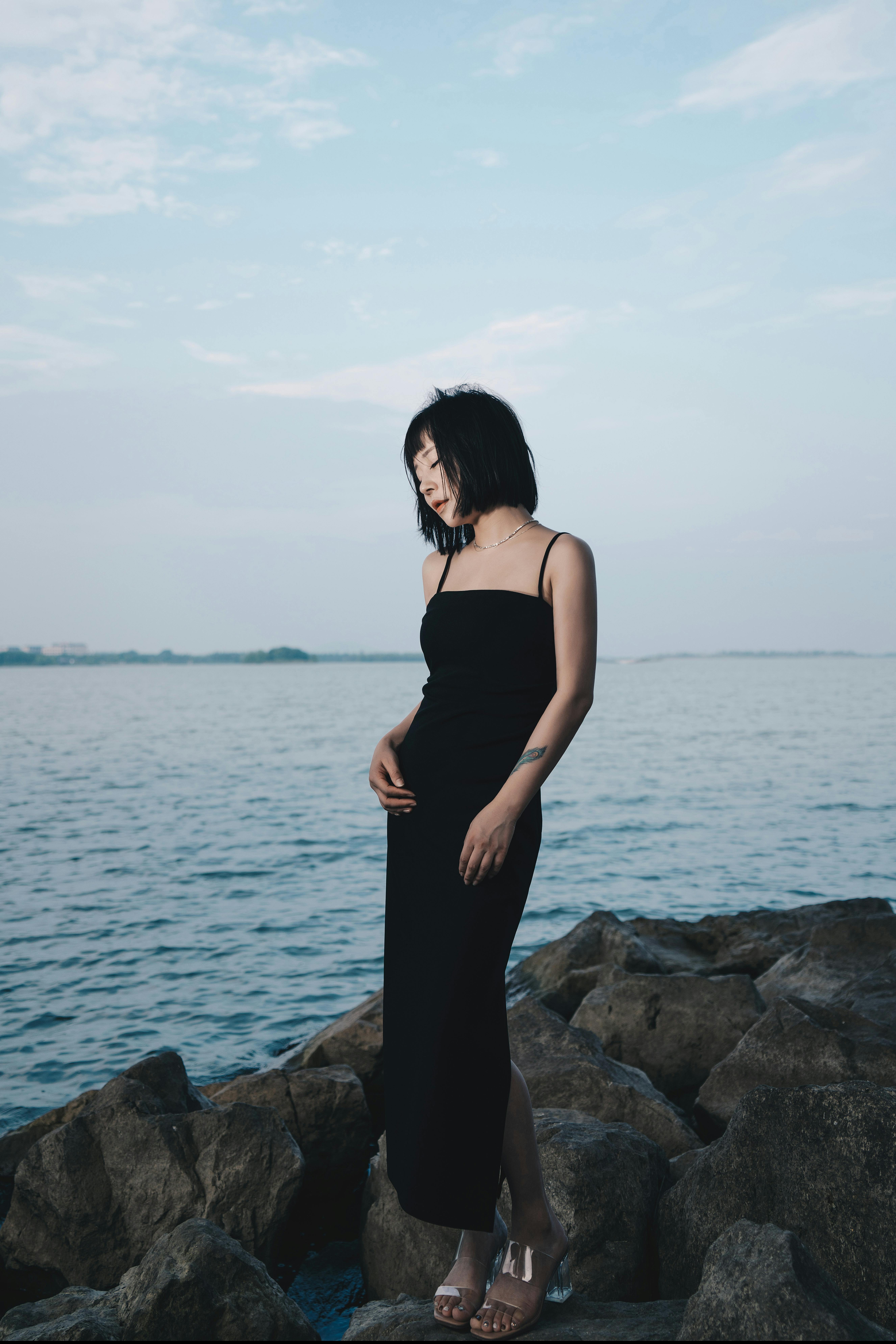 A woman in a black dress elegantly poses on rocks by the water, exuding fashion and tranquility.