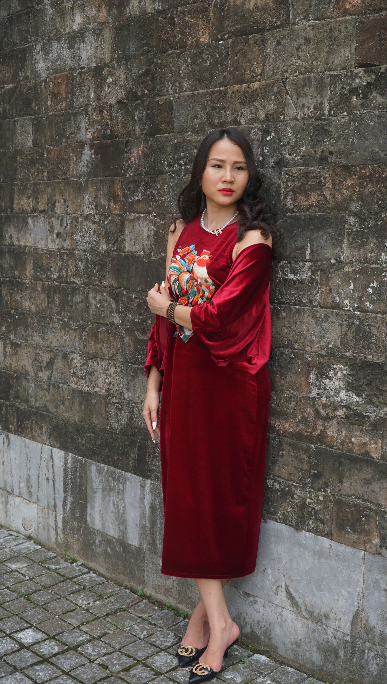 Woman in a red velvet embroidered midi dress with a pearl necklace and black pointed mules, leaning against a stone wall.