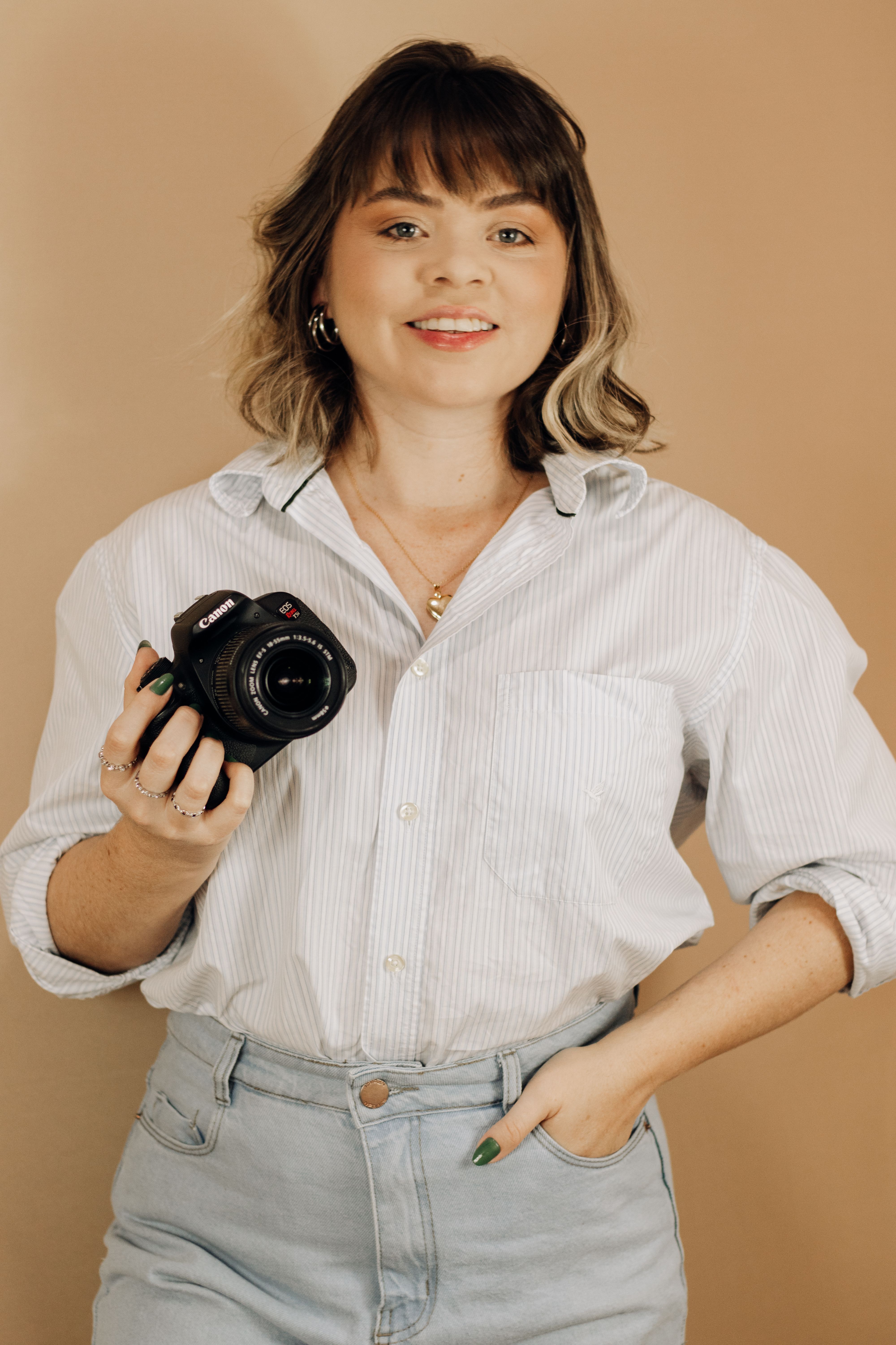 A smiling woman holding a camera, wearing a casual outfit indoors.