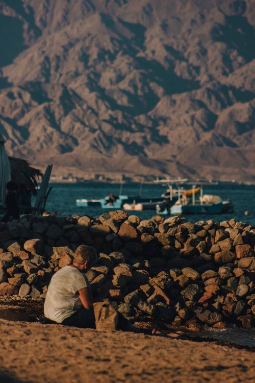 Free A woman sitting on a rocky beach in South Sinai, Egypt, enjoying the mountainous and seaside view. Stock Photo