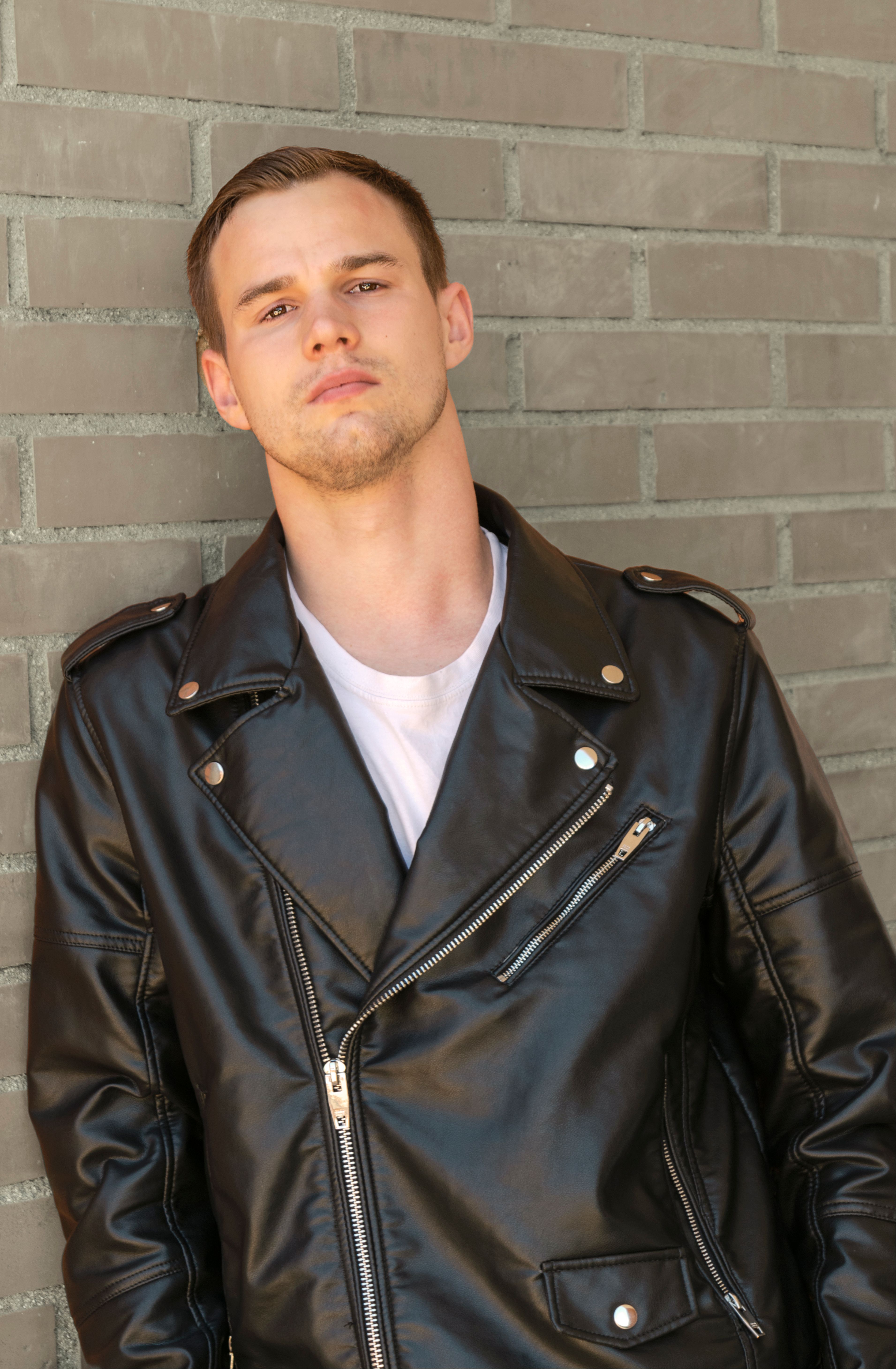 Portrait of a confident young man in a stylish black leather jacket leaning against a brick wall.