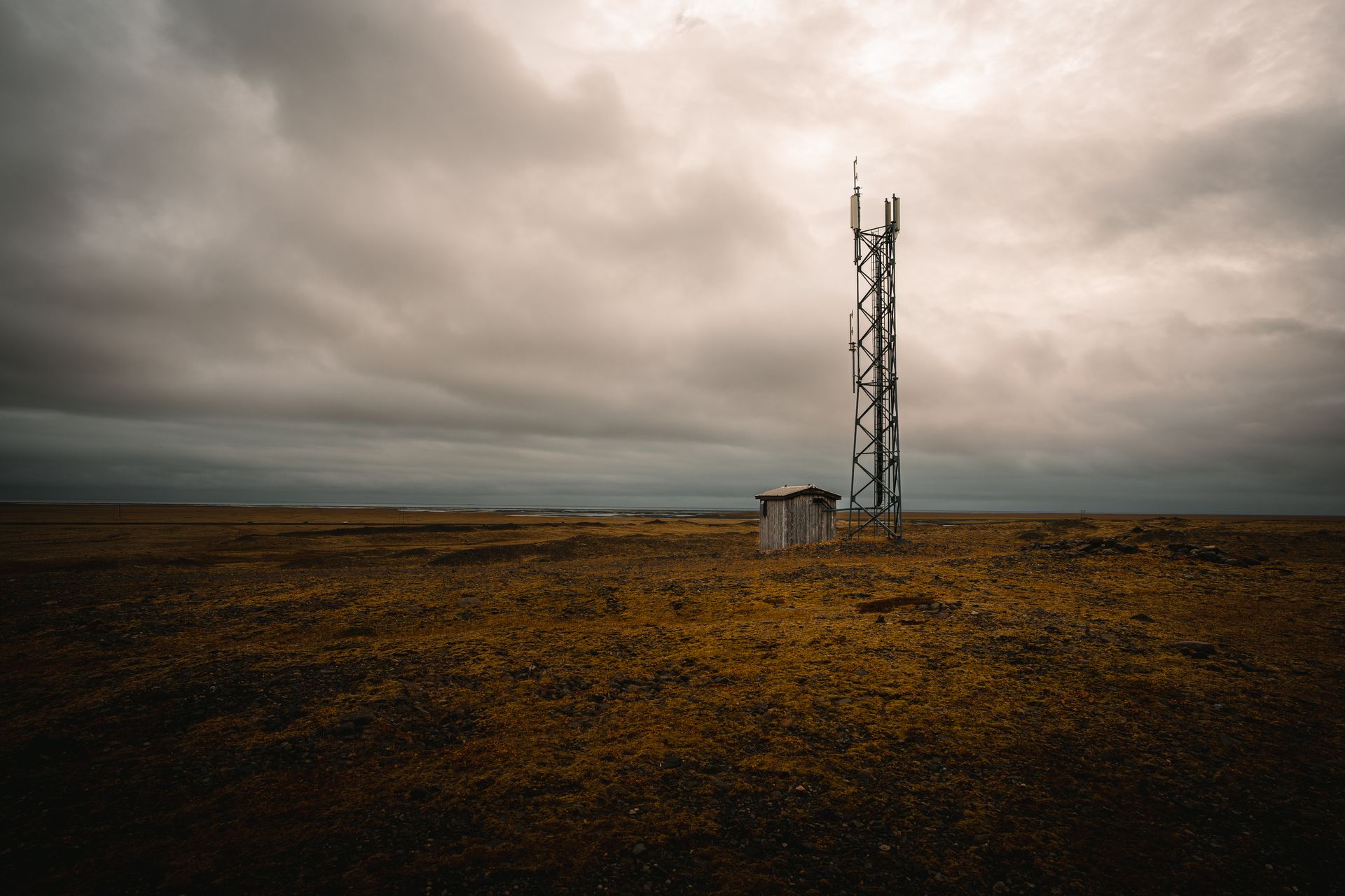 A lone relay tower and shed stand in a vast barren landscape under a dramatic overcast sky.