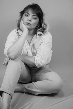 Black and white portrait of a thoughtful woman sitting barefoot in a studio.