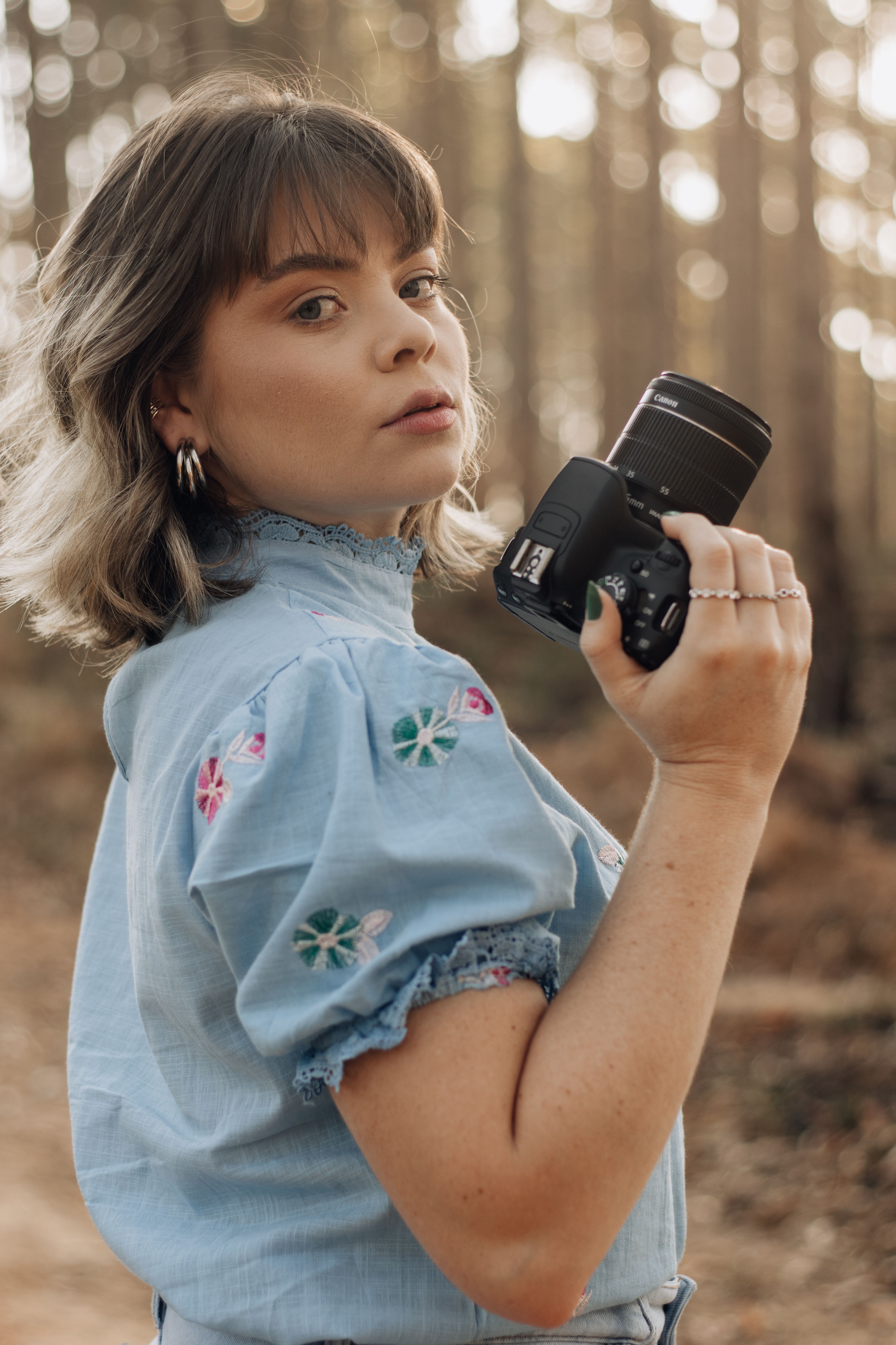 Young woman in a forest holding a camera, wearing a floral patterned blouse in São Paulo, SP, Brazil.