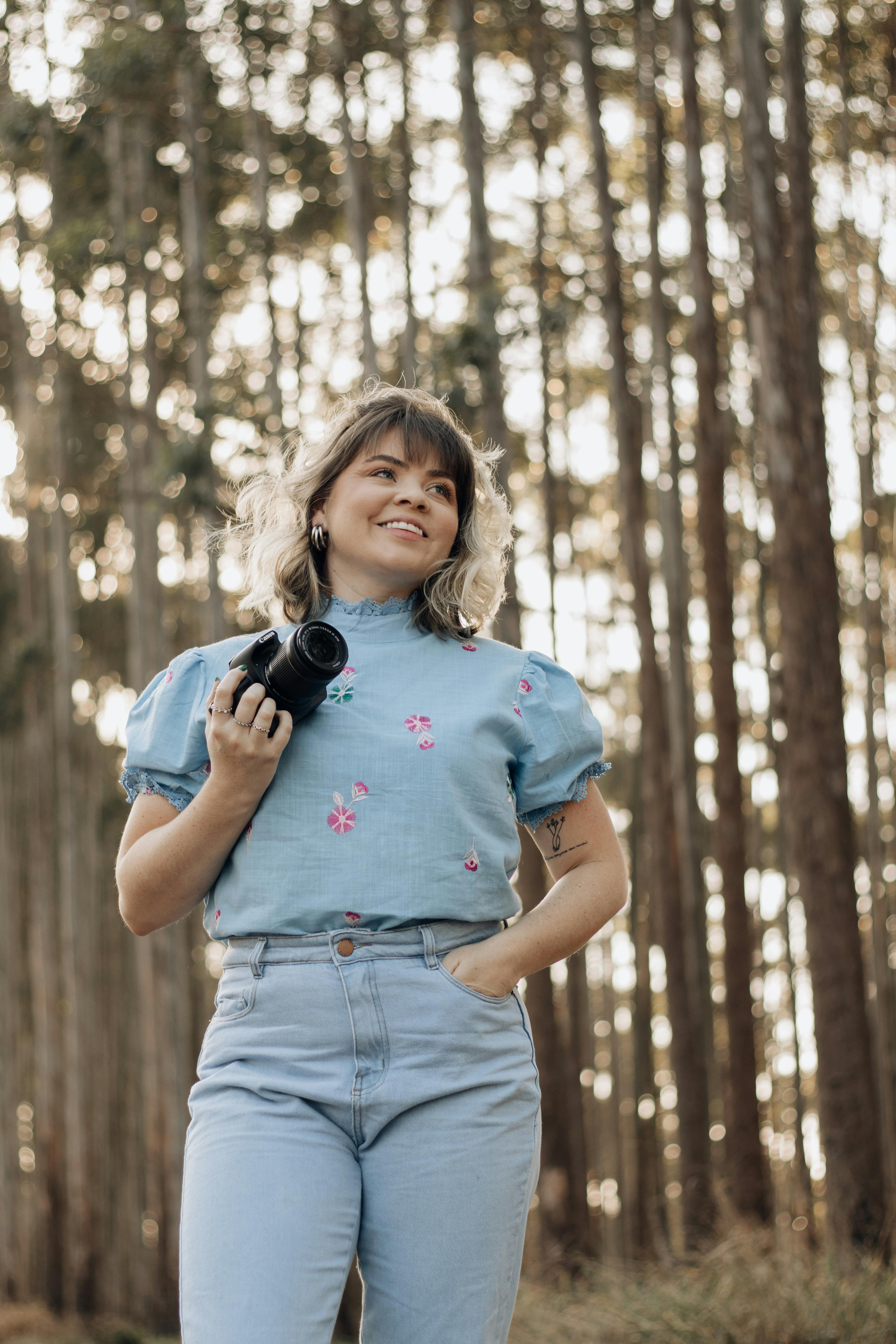 A woman in a short-sleeved blouse holding a camera, standing in a lush São Paulo forest.