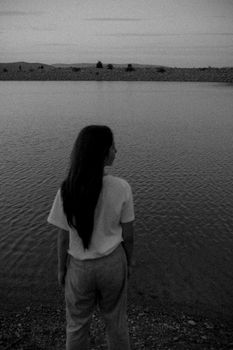 A woman stands thoughtfully by a calm river in black and white.