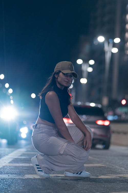Woman in Turtleneck Top and Baseball Cap Crouching on Street at Night