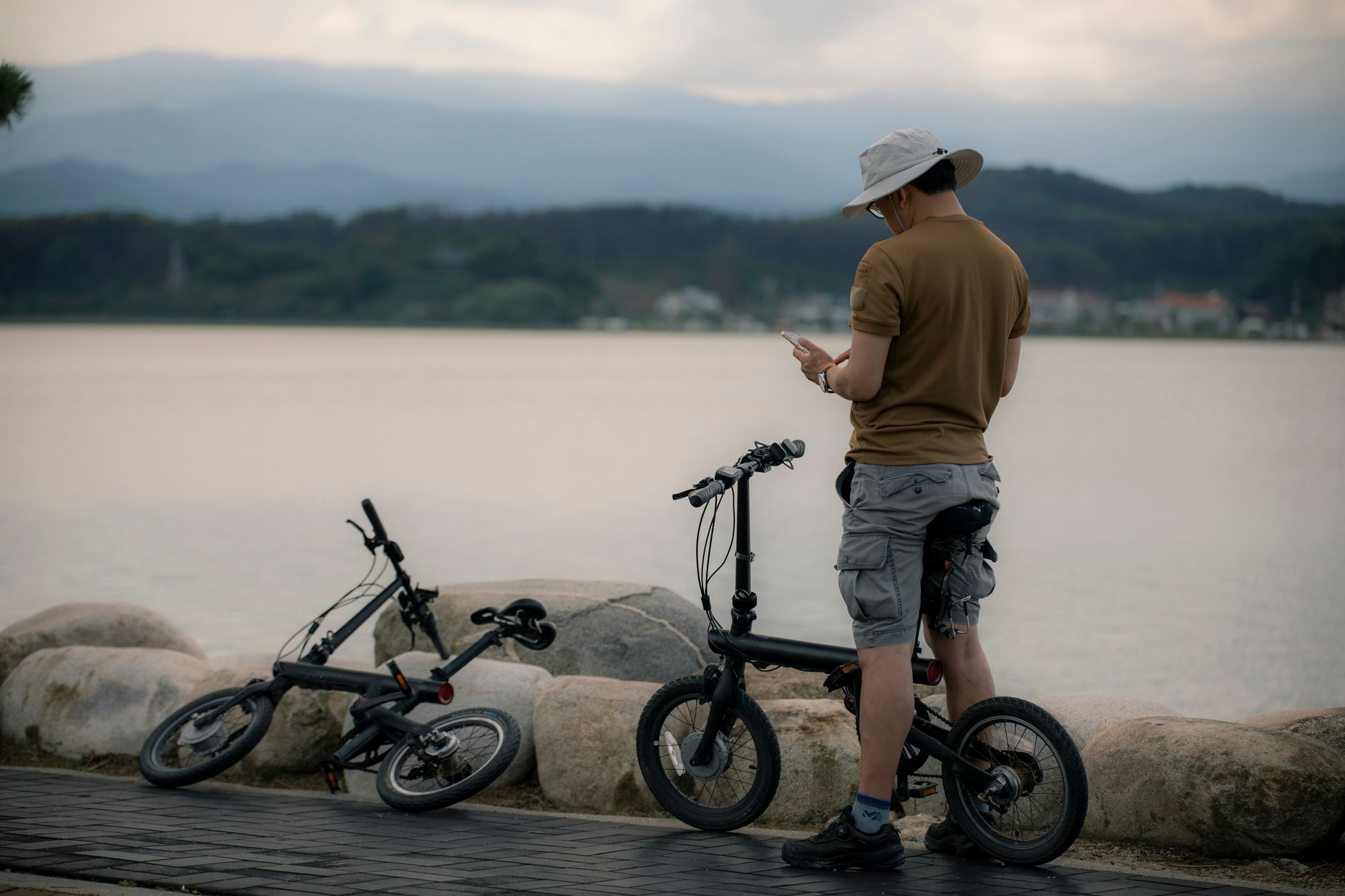 A man stands by a tranquil lake in Gangneung, South Korea, checking his smartphone beside two bicycles.