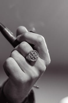 Monochrome image of a hand holding a pipe, showcasing a detailed signet ring.