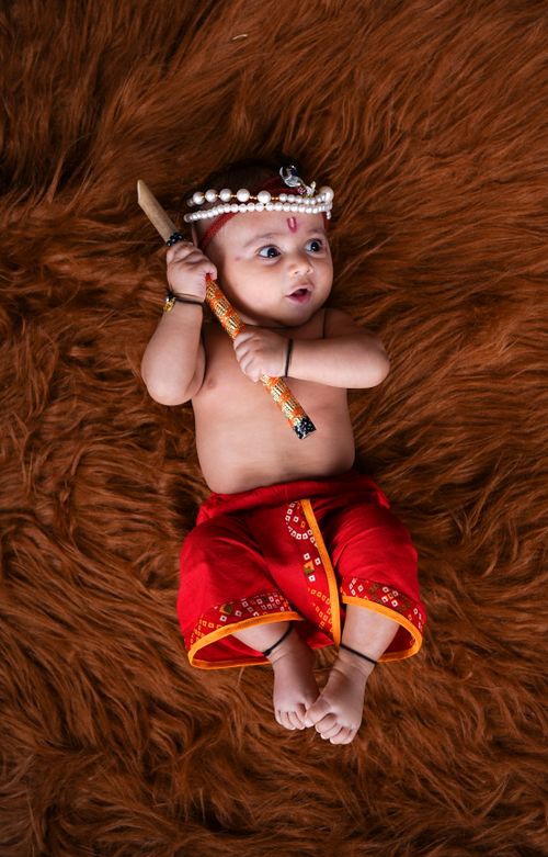Free Cute baby dressed in traditional attire holding a stick, lying on a soft brown surface. Stock Photo