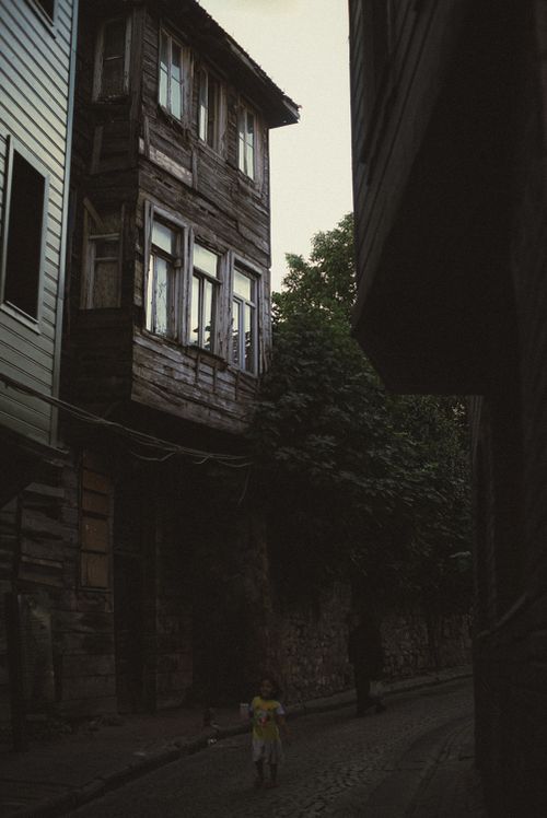 Free A child runs along a cobblestone street between old wooden houses, capturing a timeless moment in a quaint city alley. Stock Photo