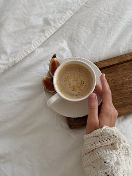 A close-up of a hand holding a coffee cup with figs on a bed, exuding warmth and comfort.