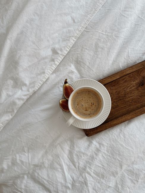 A cozy setup featuring a coffee cup with figs on a wooden tray atop a white bedspread.