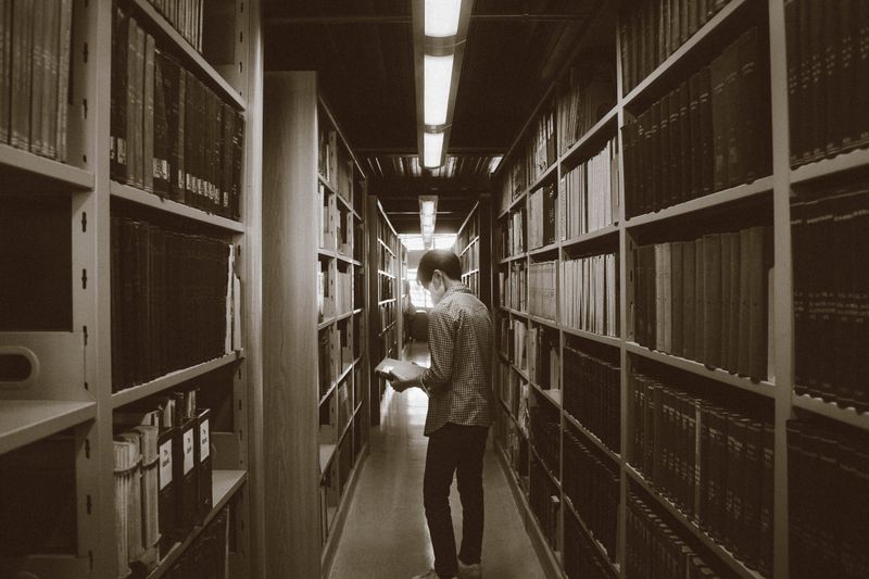 A figure reading between tall archive shelves in a dim library aisle — photo by Roman Castillo on Pexels