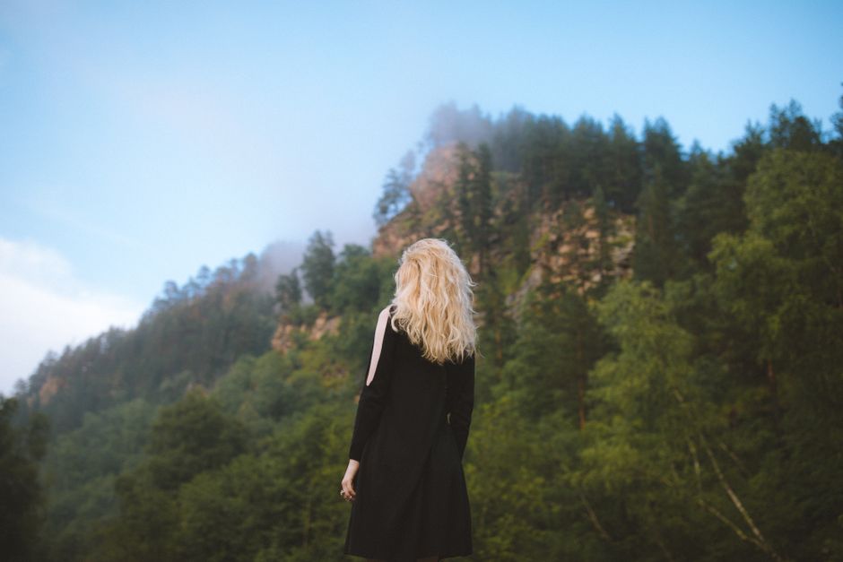 A blonde woman in a black dress stands in a misty mountain forest, capturing tranquility and adventure.