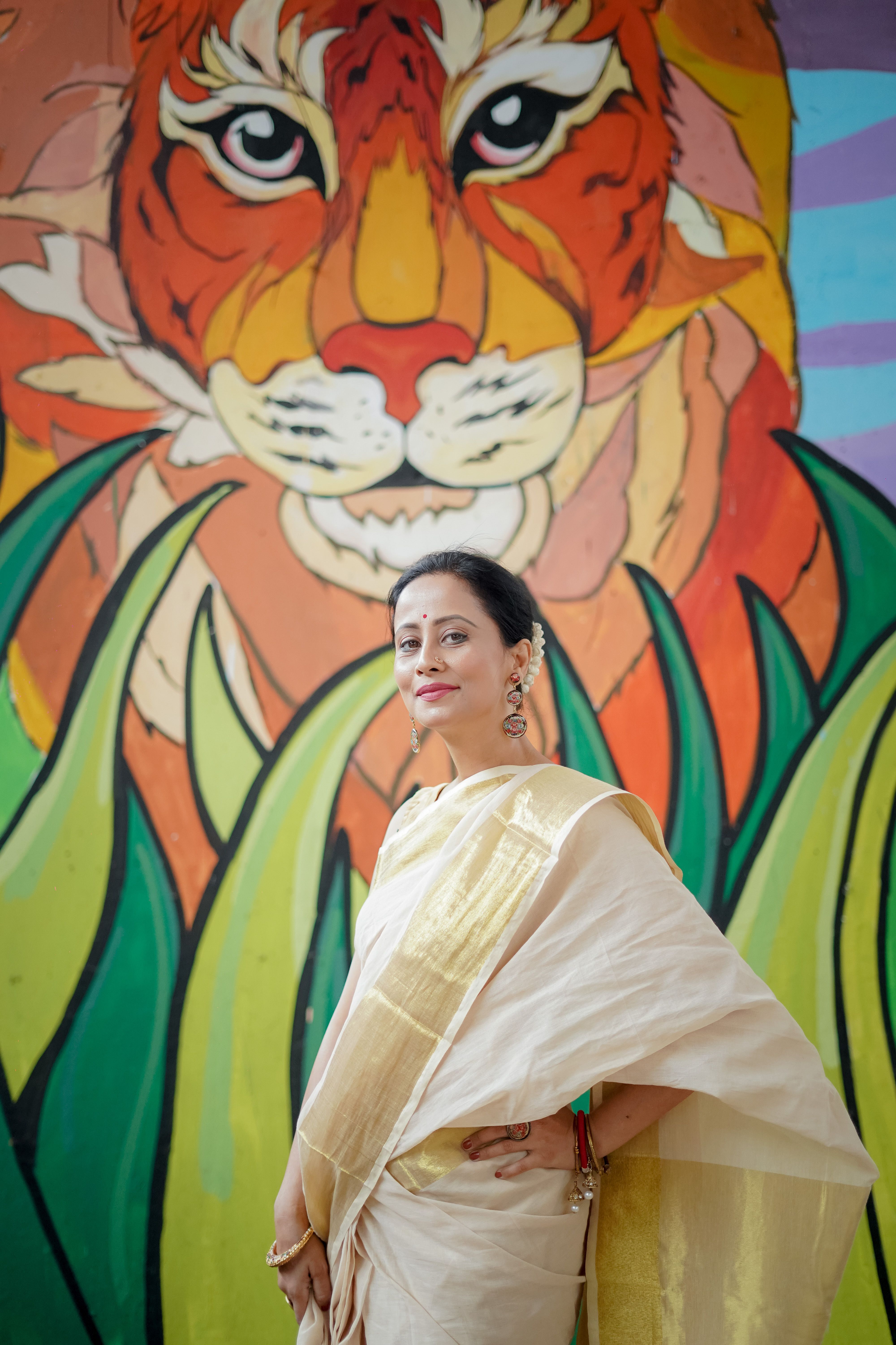 Stylish woman in a beige saree poses with a colorful tiger mural as a backdrop.