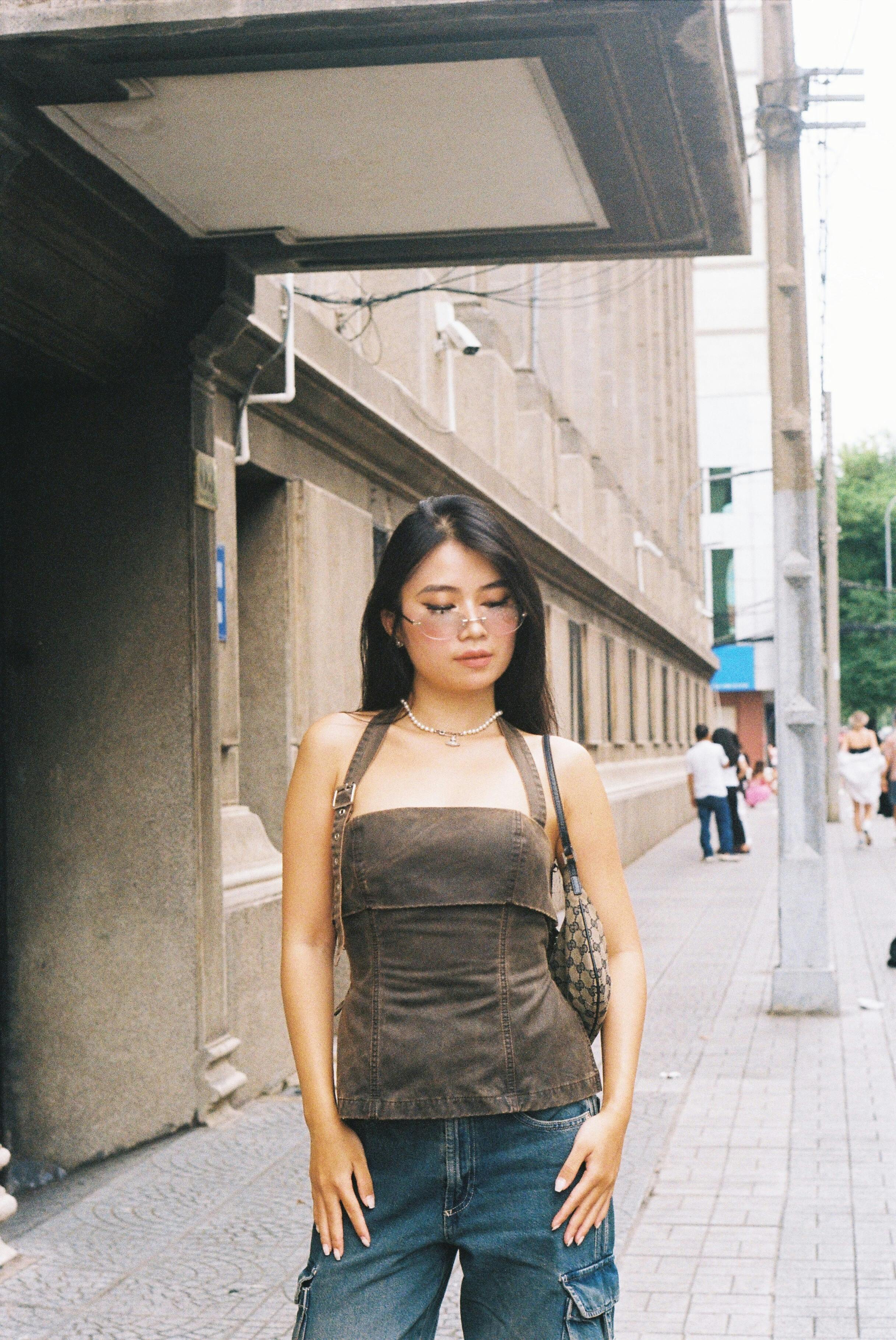 Fashionable young woman in brown leather top standing confidently on a city sidewalk.