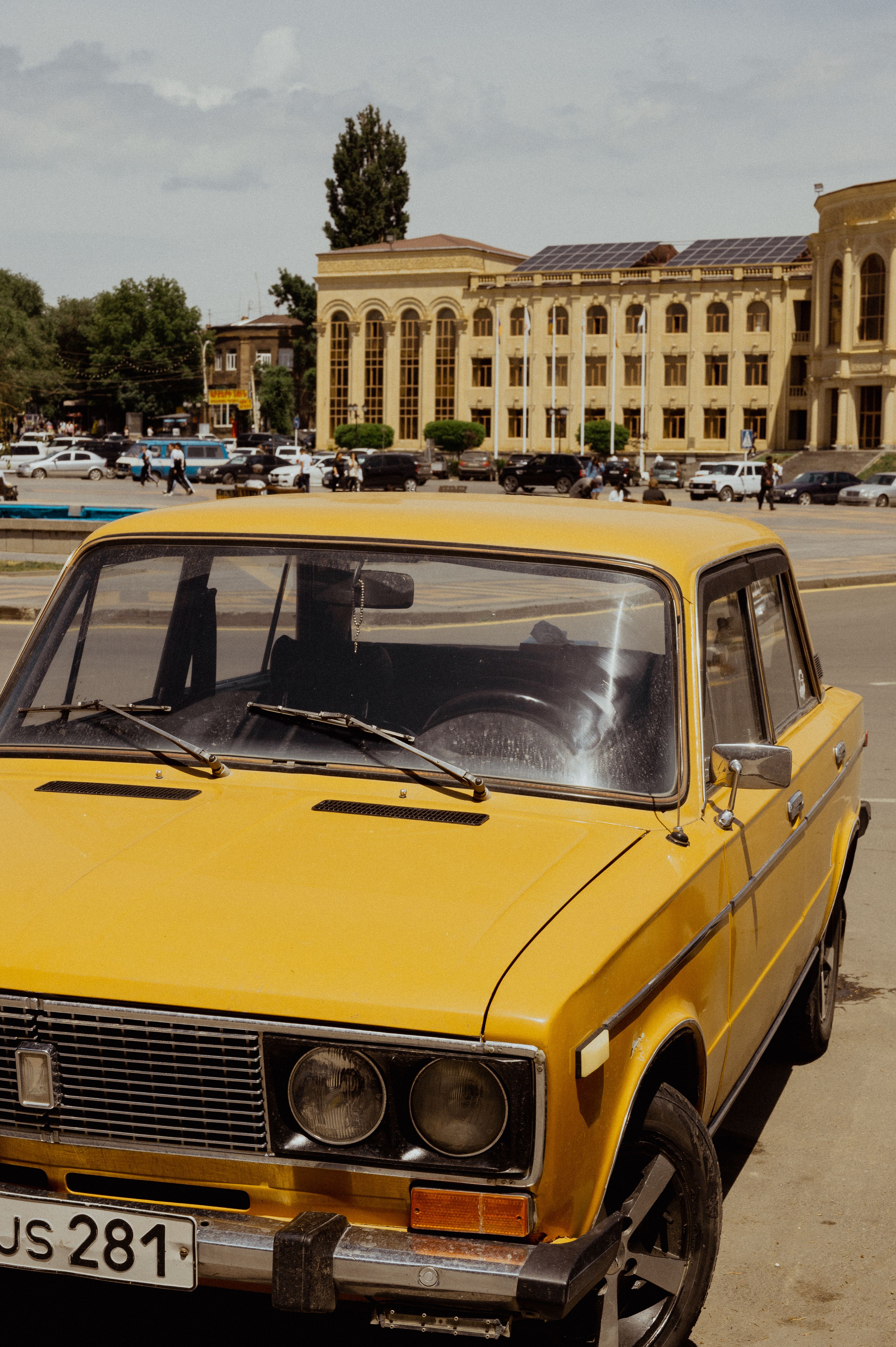 A vintage yellow car parked in a city street with historic architecture in the background.