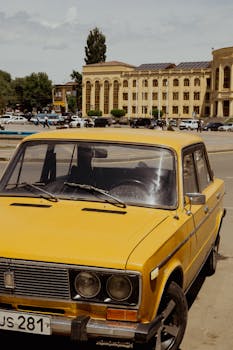 A vintage yellow car parked in a city street with historic architecture in the background.