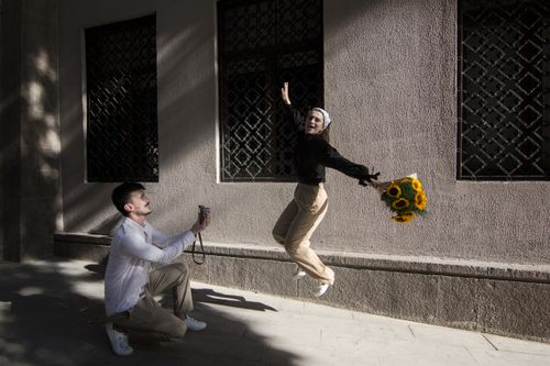 Free A woman jumps joyfully with sunflowers while a man photographs her against an urban backdrop. Stock Photo