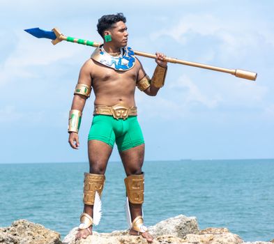 A shirtless man in a superhero warrior costume stands on rocks by the sea holding a spear.