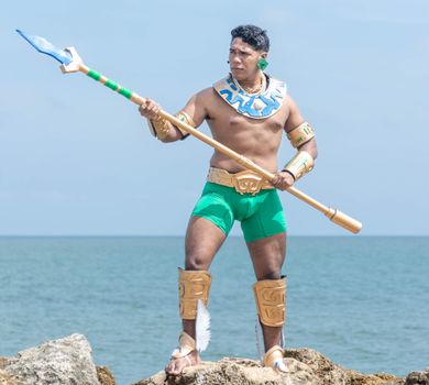 A man in tribal cosplay with a spear stands on rocks by the ocean, embodying a warrior spirit.
