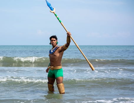 A man dressed as a warrior in green poses with a spear in the ocean waves.