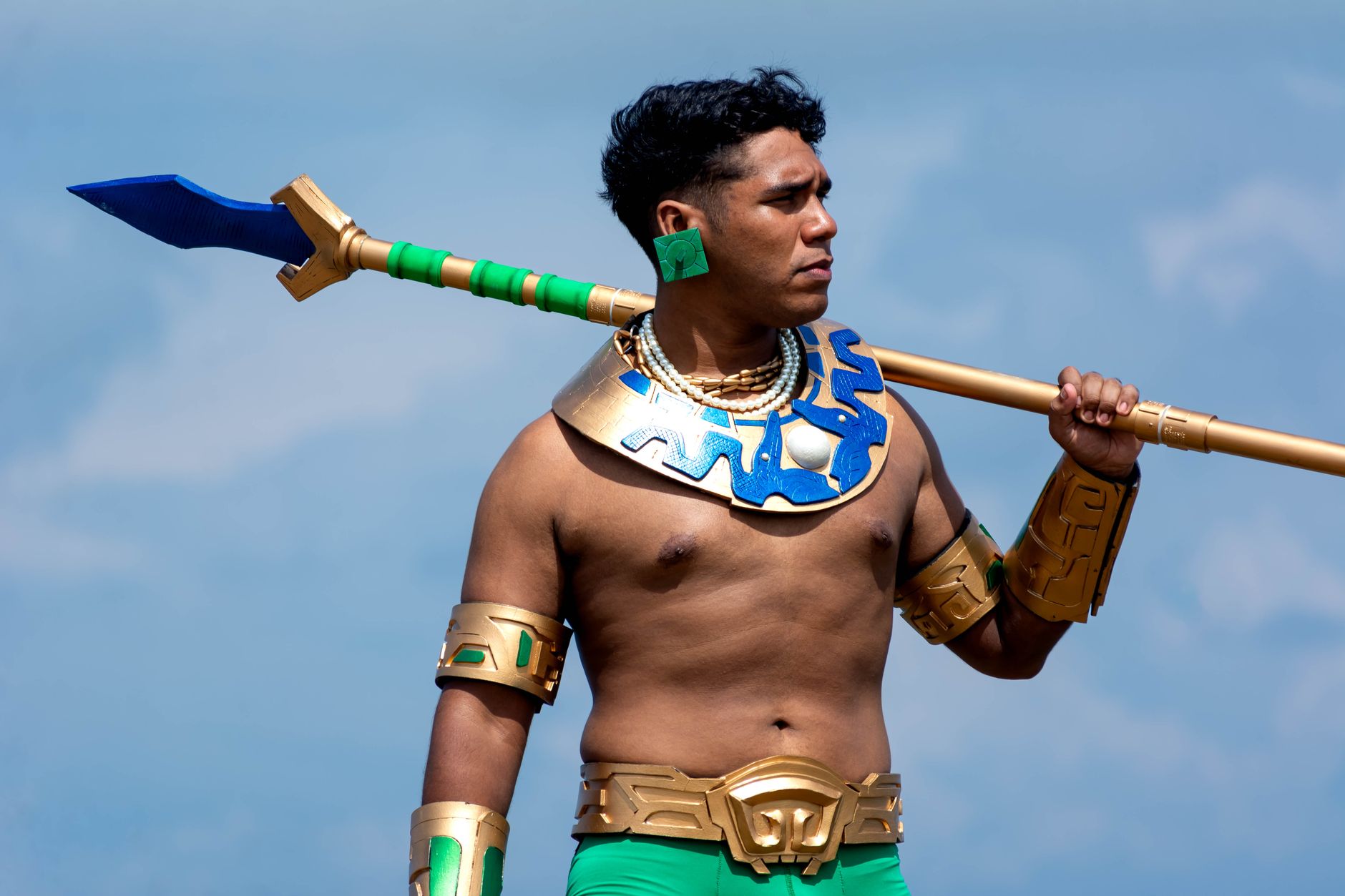 A muscular man in warrior costume standing outdoors, holding a spear against a blue sky.