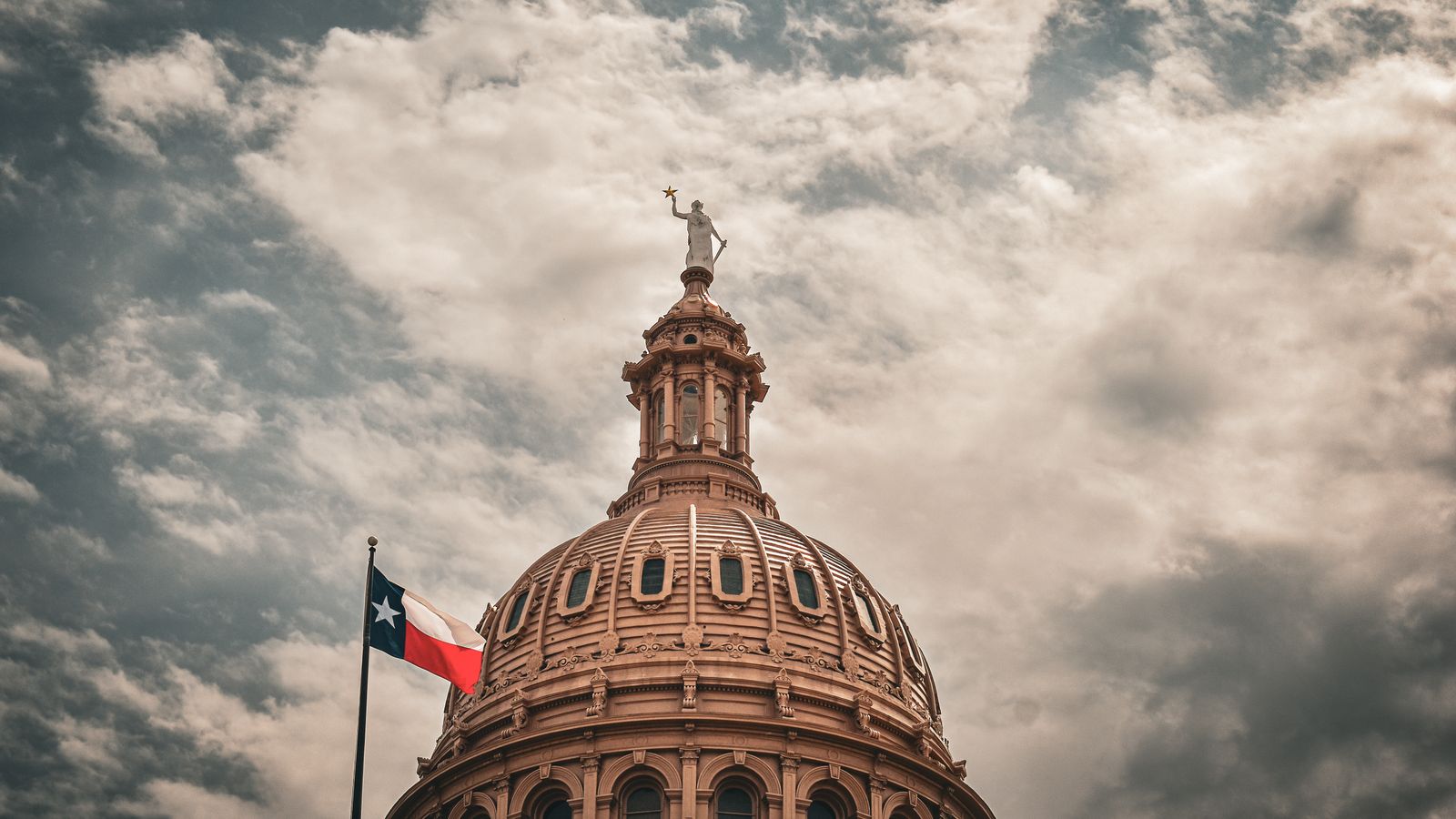The dome of the Texas State Capitol with a Texas flag under a cloudy sky.