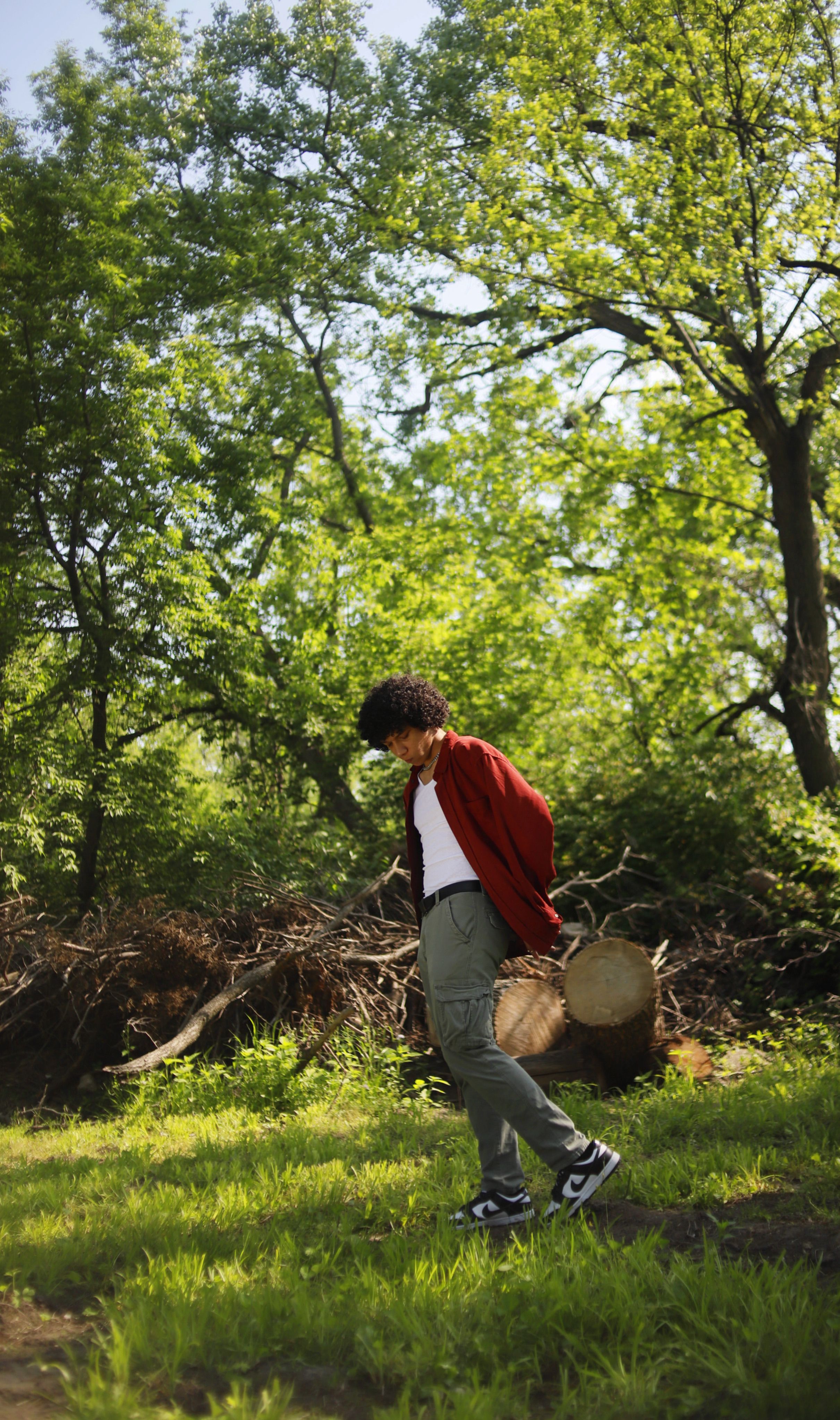 A man with curly hair walks through a vibrant park surrounded by trees and grass.