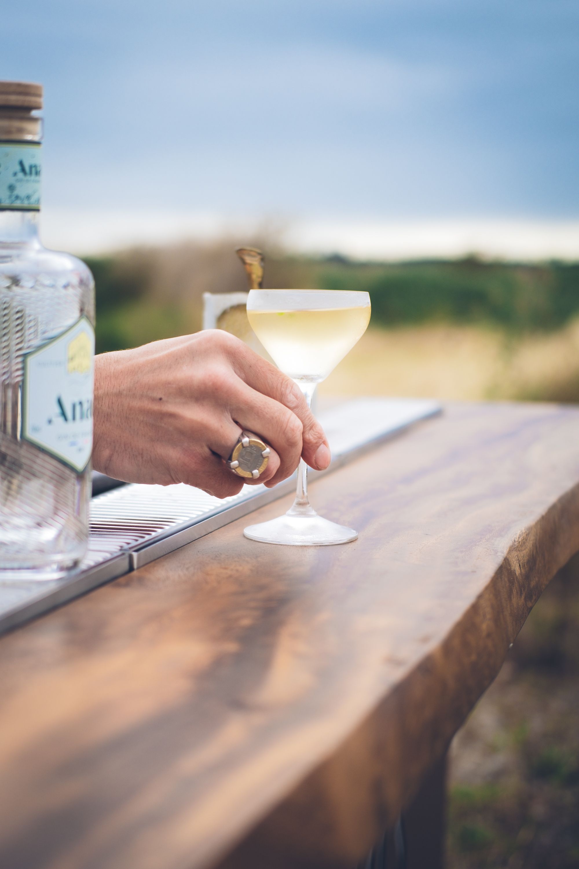 Close-up of a hand holding a cocktail glass on a wooden bar counter outdoors.