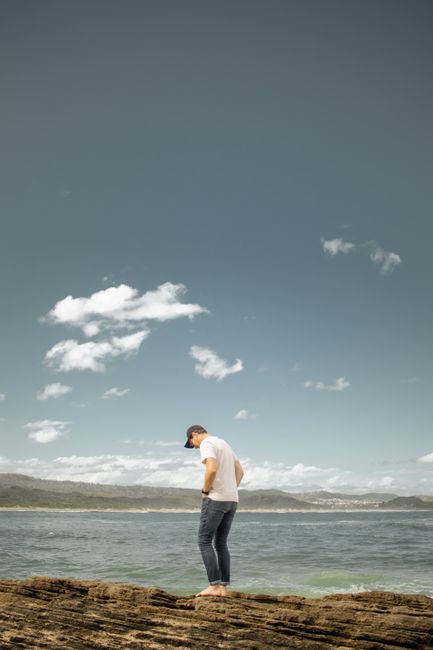 A man stands on a rocky shore, looking contemplative by the sea under a bright sky.