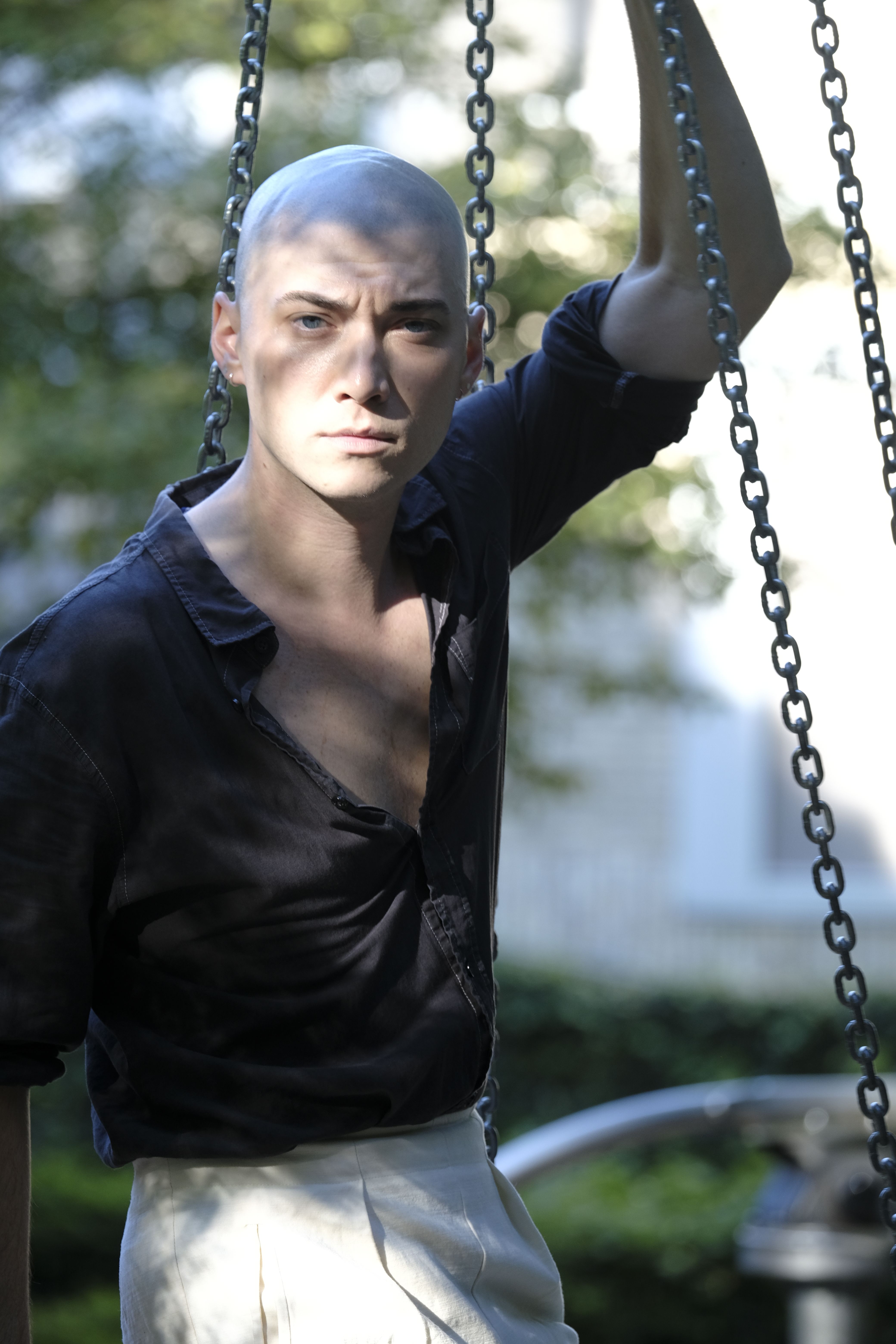 Bald man posing confidently on a playground swing, wearing dark shirt.