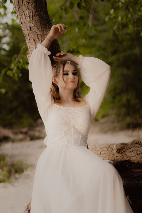 Free A serene bridal portrait featuring a woman in a flowing white dress posing against a tree in nature. Stock Photo