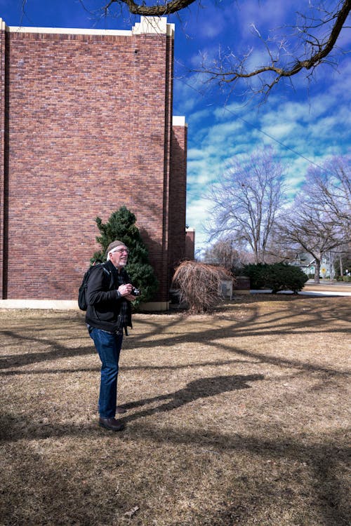 Elderly Man with a Camera Standing on the Grass in front of a Building 