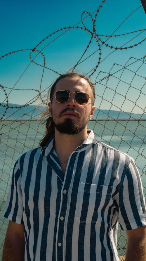 A Man in a Shirt and Sunglasses Standing in front of a Fence with a Barbed Wire
